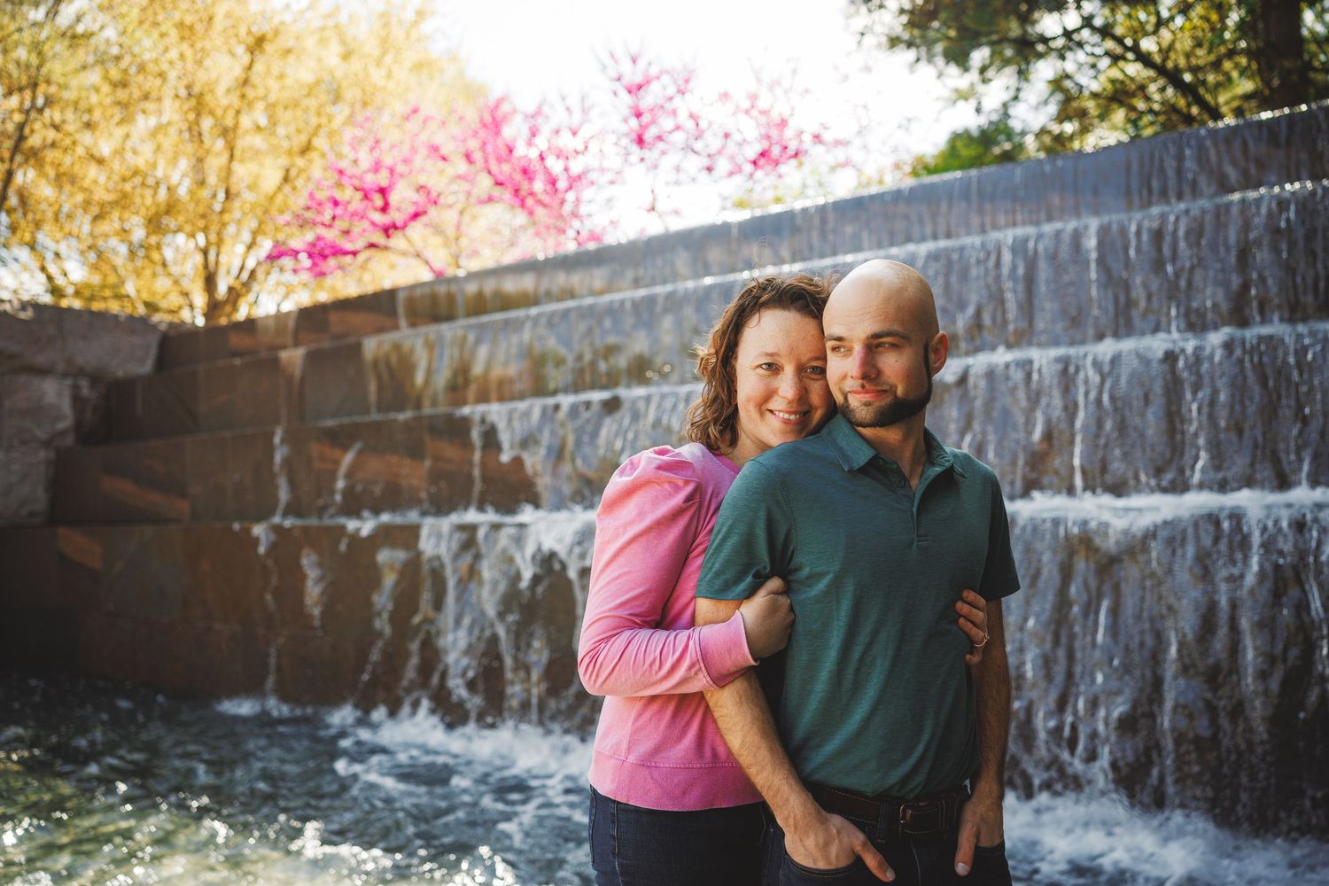 Couple embracing in front of a cascading stone waterfall with blooming trees in the background.