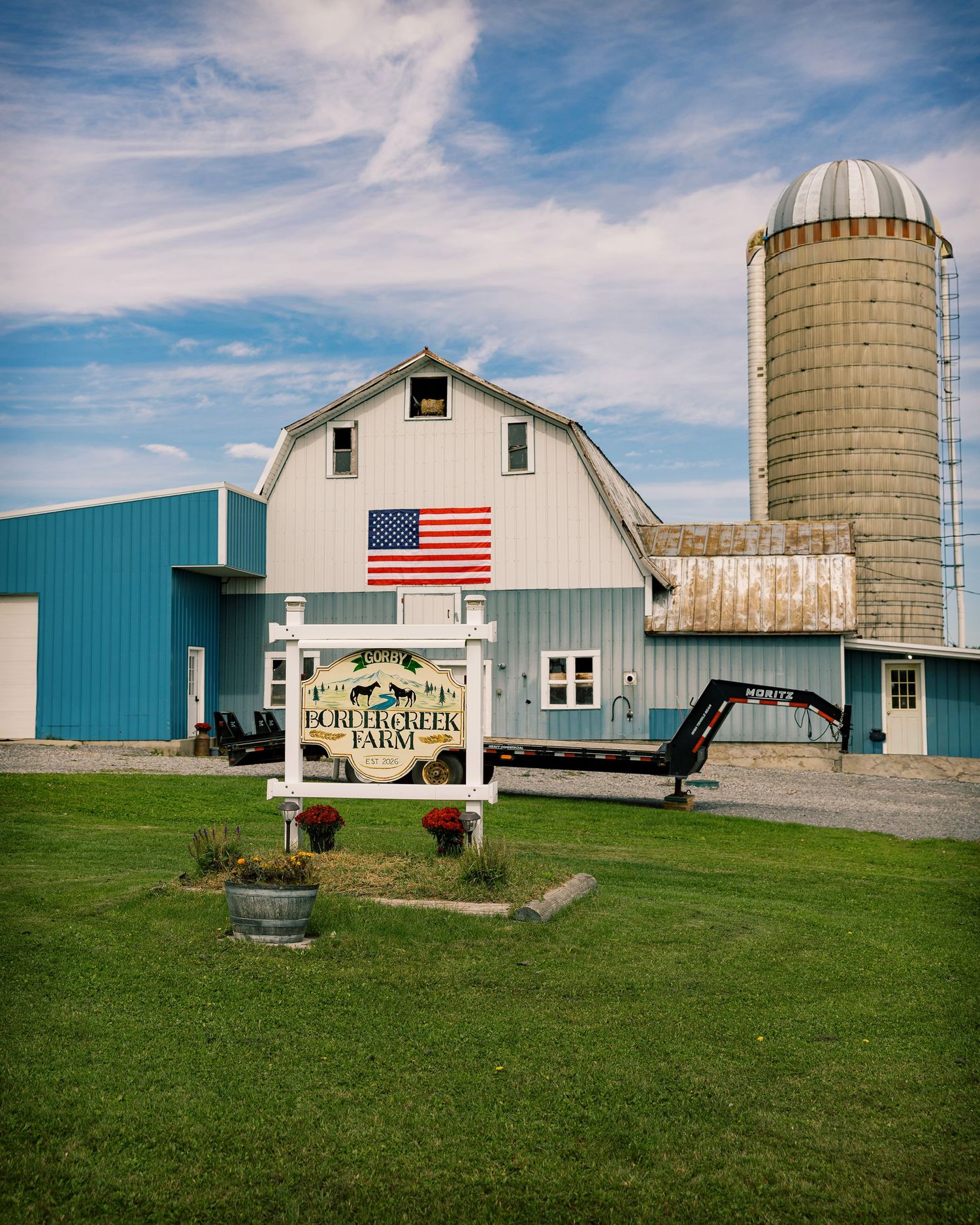White and blue farm barn with an American flag, a tall stone silo, and a welcome sign on a green lawn under a blue sky.