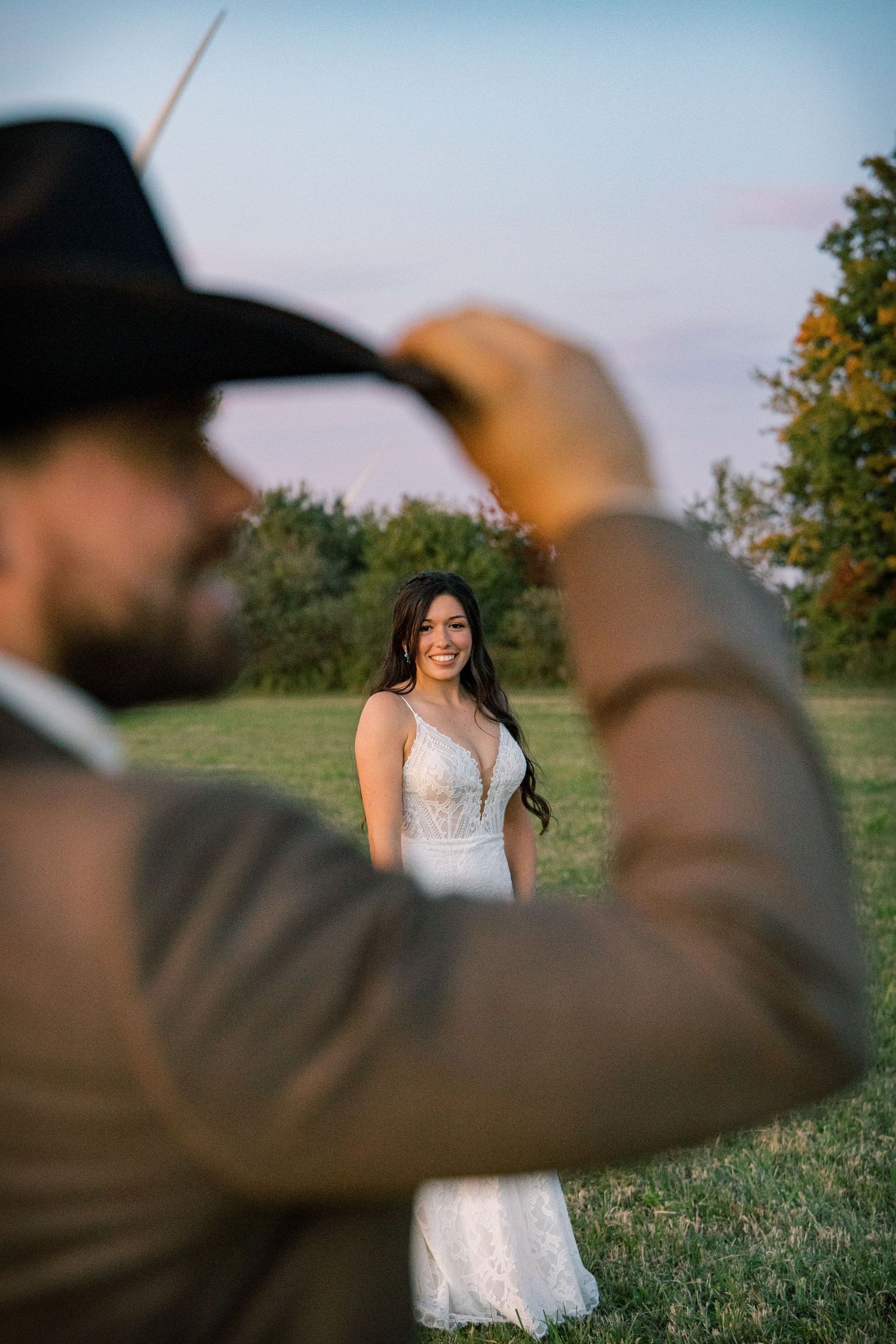 Smiling bride in a white lace wedding dress stands in a grassy field at sunset while a bearded groom in a black cowboy hat tips his brim.