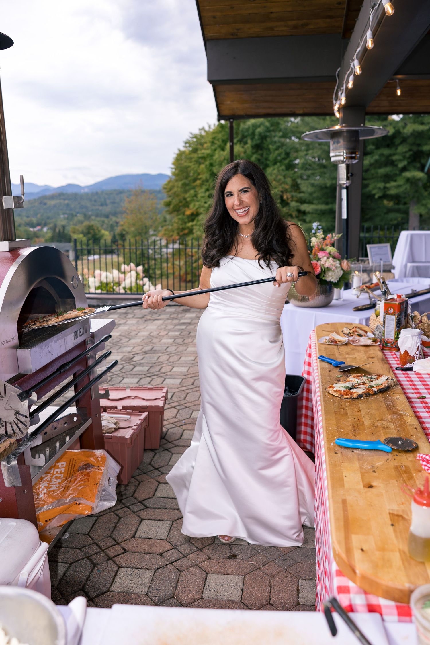 Bride in white gown using a pizza peel at an outdoor wood-fired oven during a wedding reception.