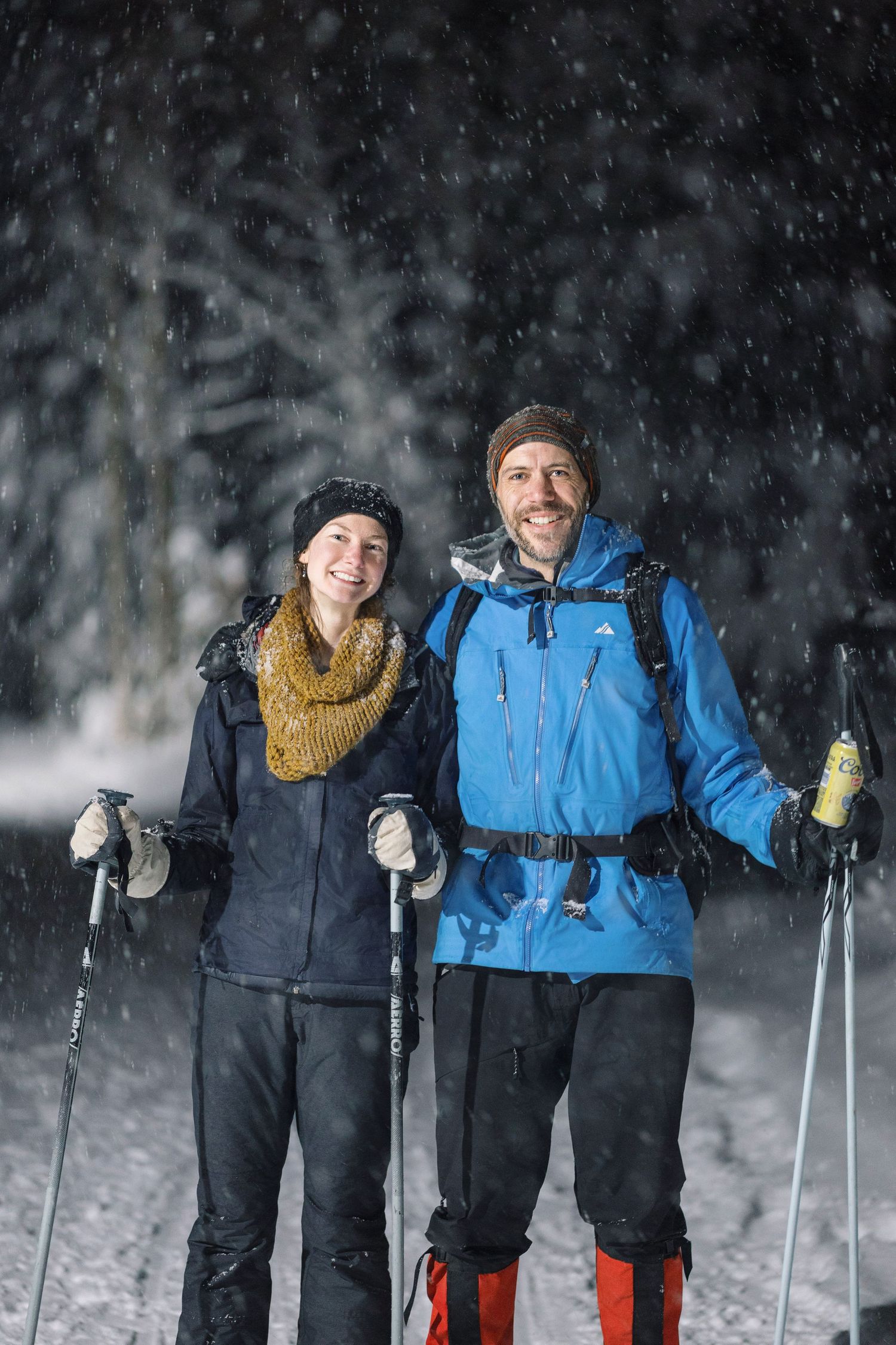 Two winter hikers with ski poles and warm clothing stand together in falling snow on a snowy trail.