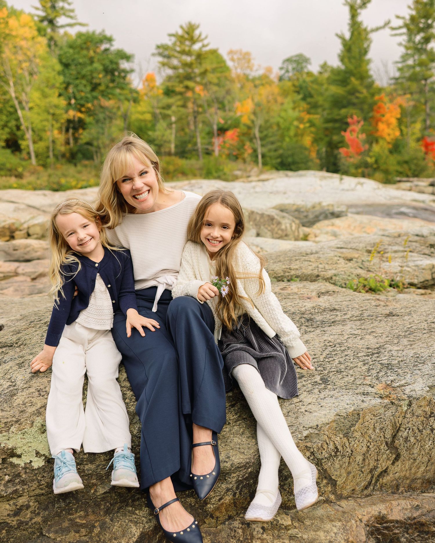 Children sit together on a large rocky outcrop surrounded by autumn trees in a natural outdoor setting.