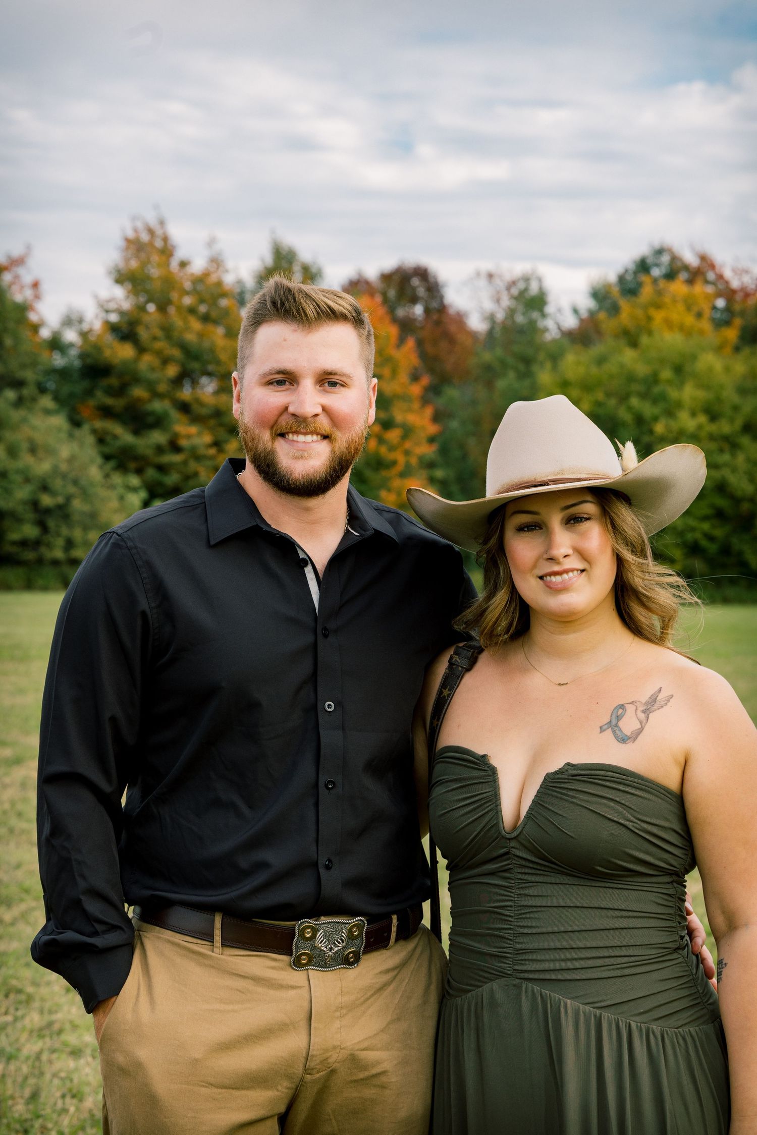 Smiling couple pose outdoors in a field with autumn trees, he in a black shirt and belt buckle, she in a green dress and cowboy hat.