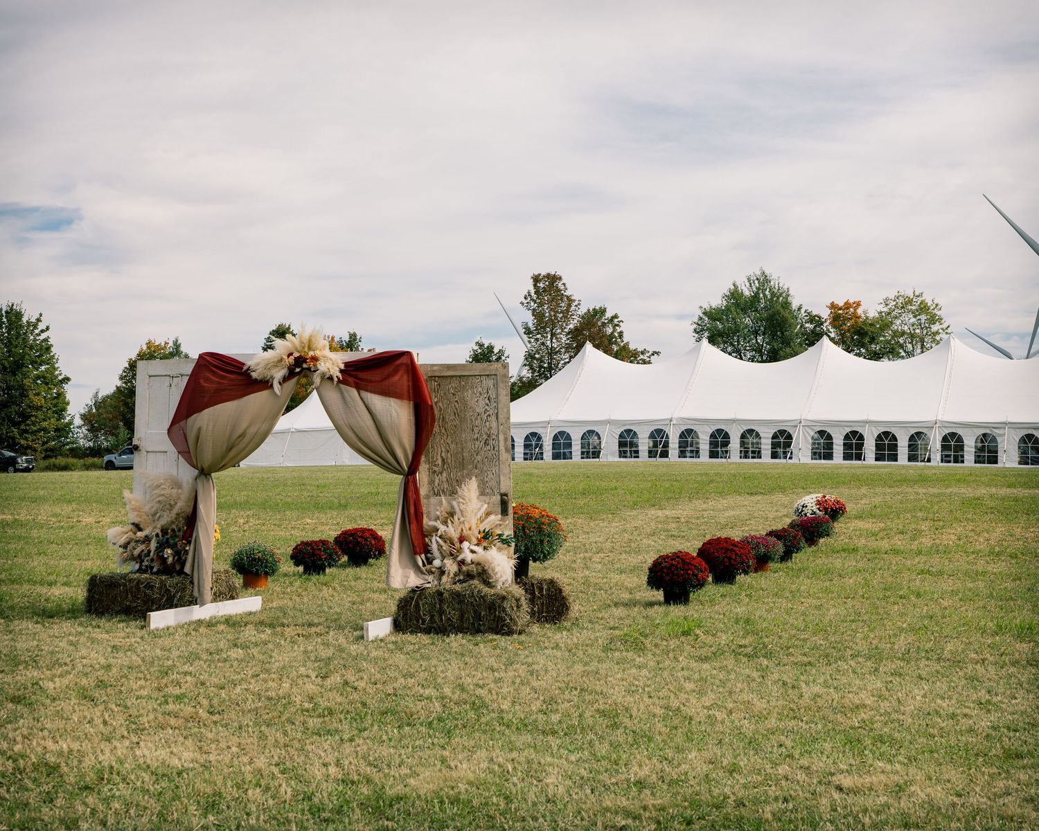 Outdoor wedding ceremony arch draped in burgundy fabric with pampas grass and mums, set on a lawn before a large white event tent.