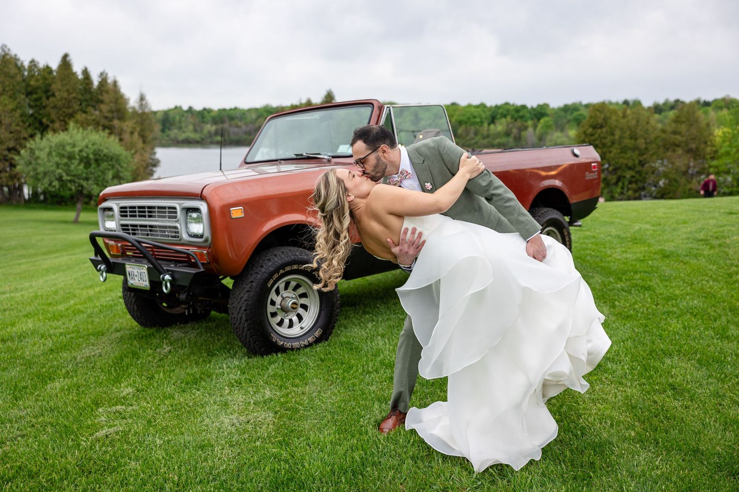 Bride and groom share a romantic dip kiss in front of a vintage orange truck on a lush green lawn.