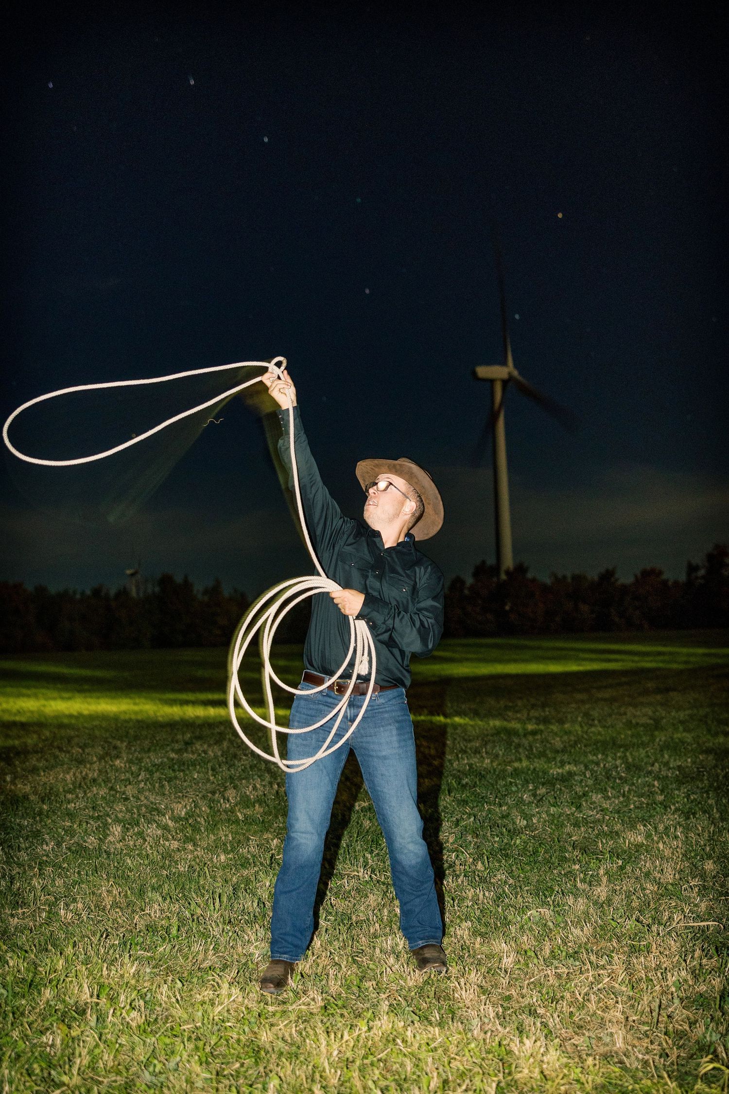 Man in a cowboy hat swinging a lasso in a grassy field at night with a wind turbine and starry sky behind him.