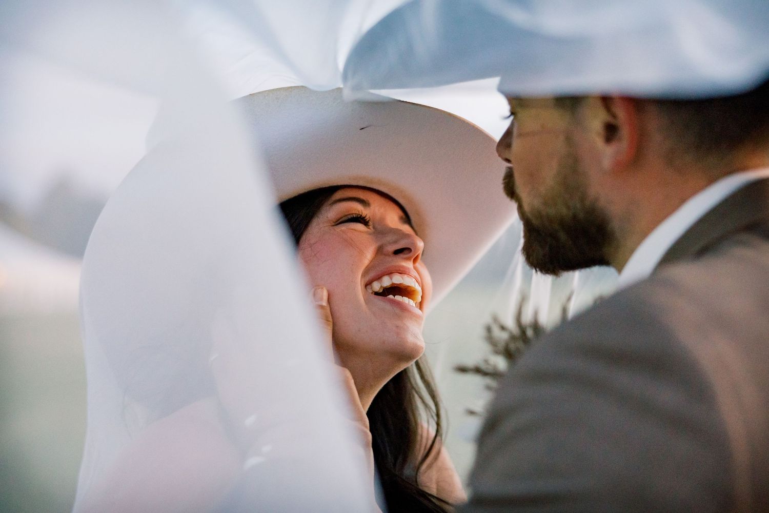 Smiling bride in a wide-brimmed white hat laughs joyfully under a sheer veil while looking up at a bearded groom in a tweed jacket.