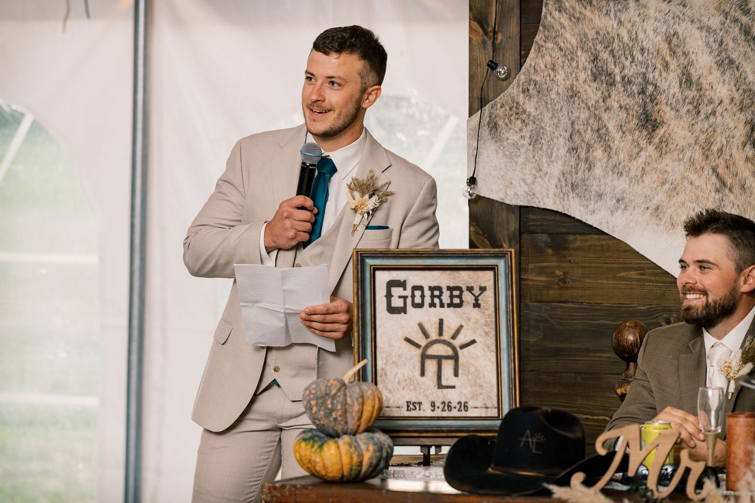 A groomsman in a tan suit speaks into a microphone beside a framed Gorby sign and pumpkins at a rustic wedding reception.
