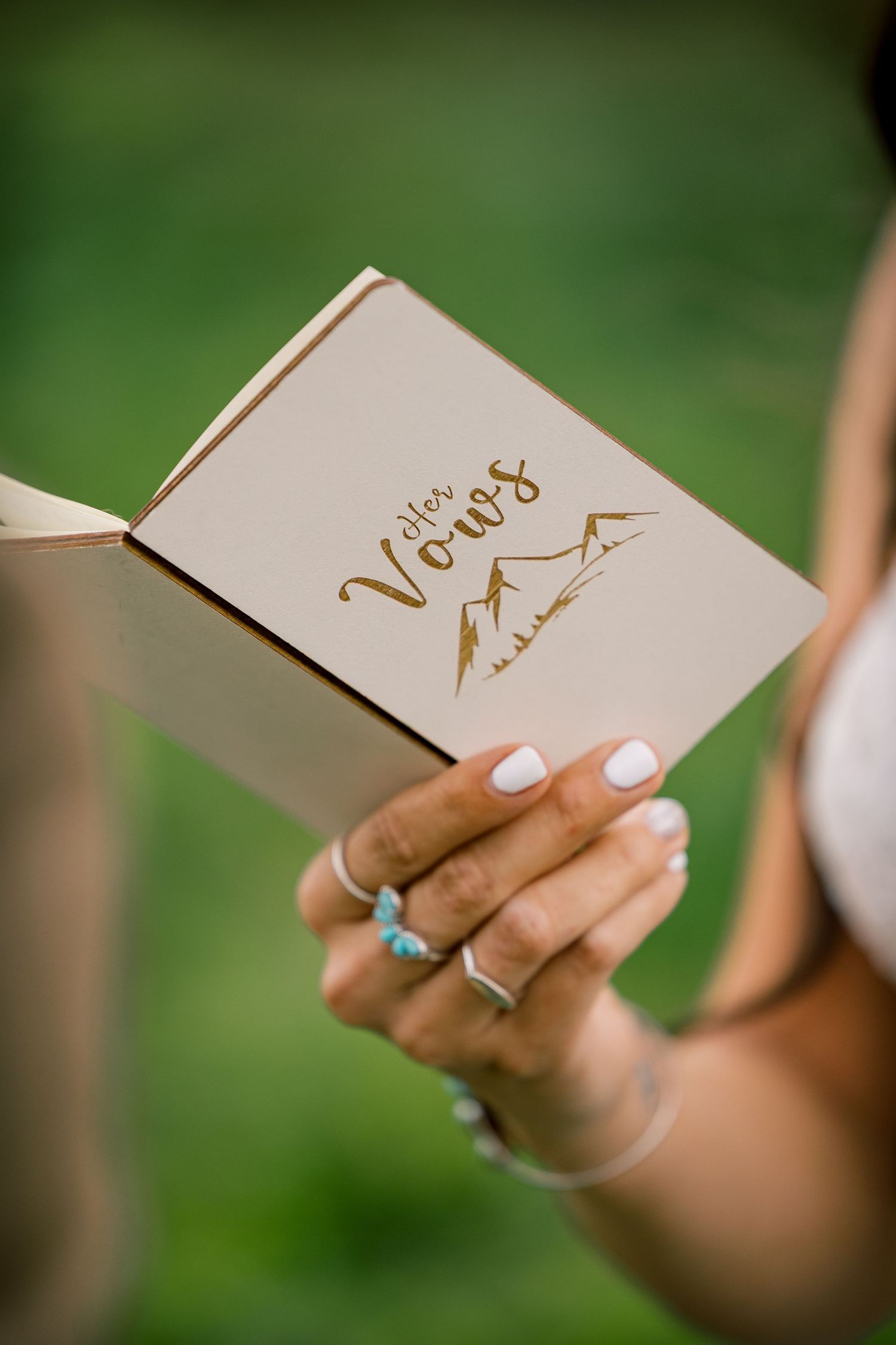 Bride's hand holding an open wedding vow book with gold mountain design and the title Her Vows, wearing turquoise and silver rings.