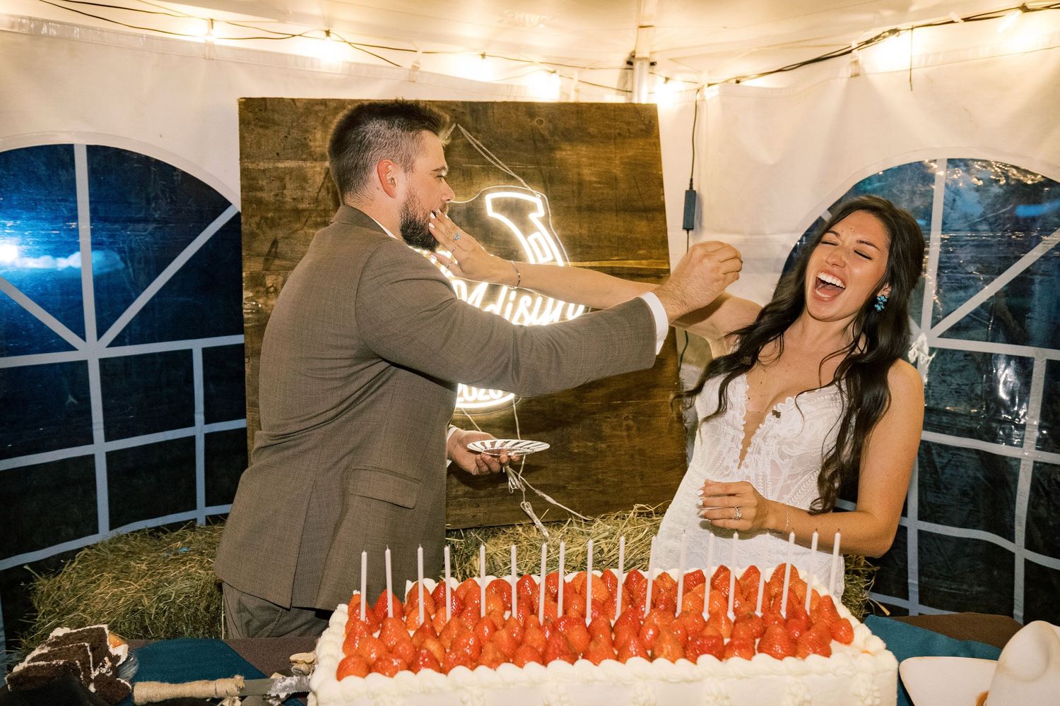 Laughing bride in a white dress and groom in a gray suit playfully smear cake at their wedding reception beside a strawberry-topped cake.