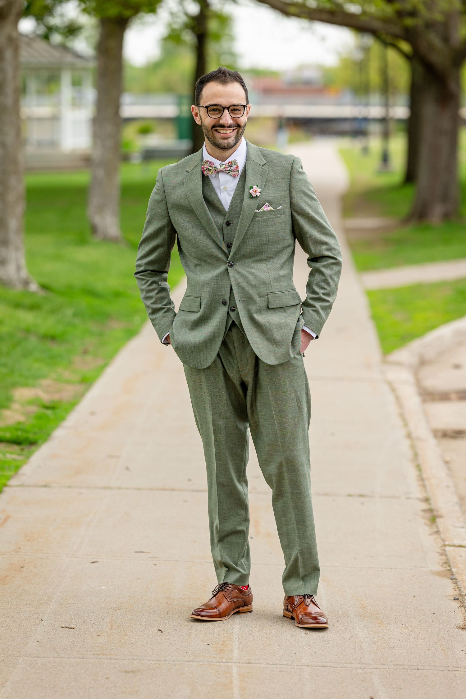Man in sage green three-piece suit with bow tie and brown oxford shoes smiling on a park pathway.