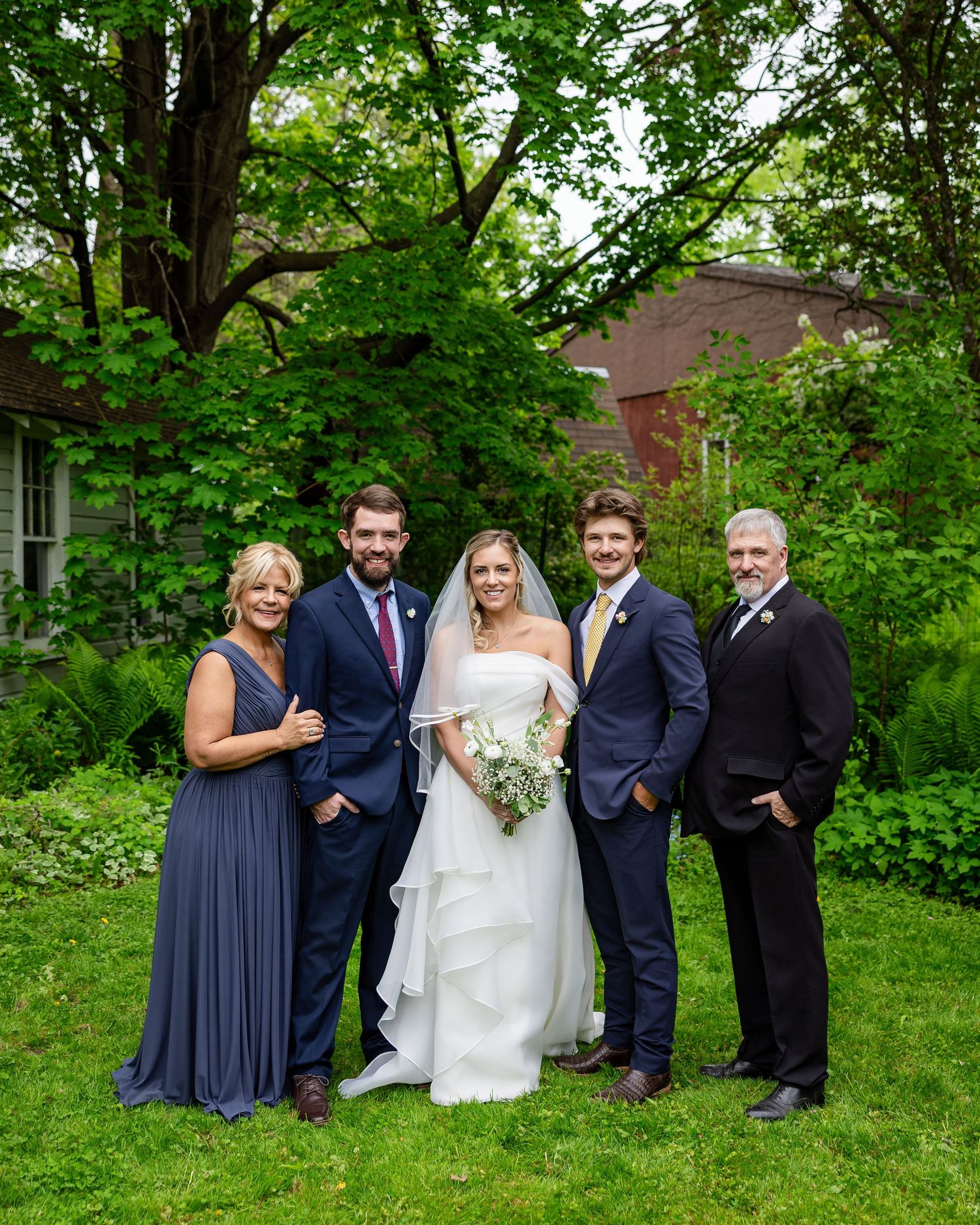 Wedding party of five posing outdoors in lush garden, bride in white gown with veil, others in navy formal attire.