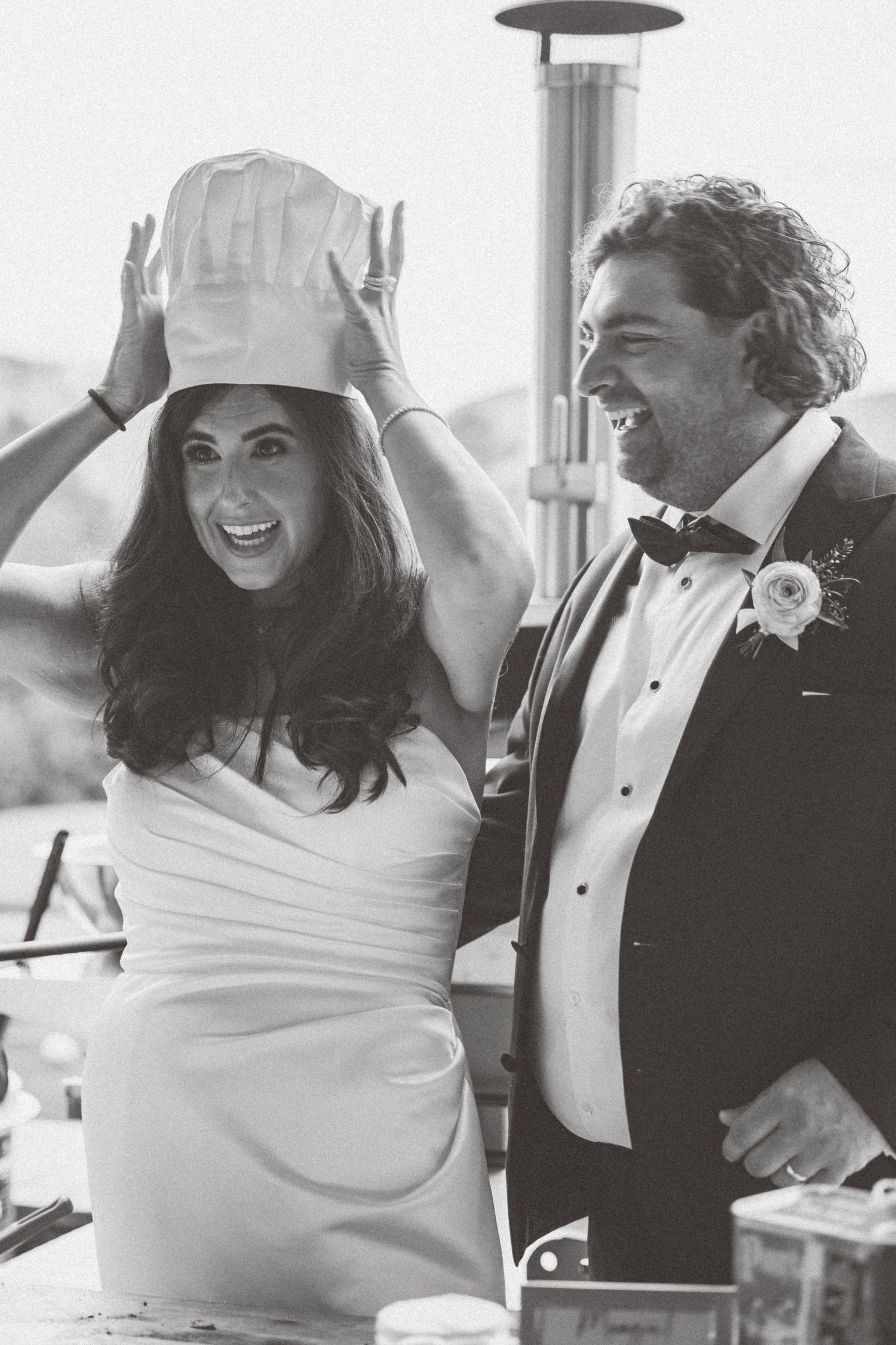 Black and white photo of a bride wearing a chef hat laughing with a groom in a tuxedo at their wedding reception.