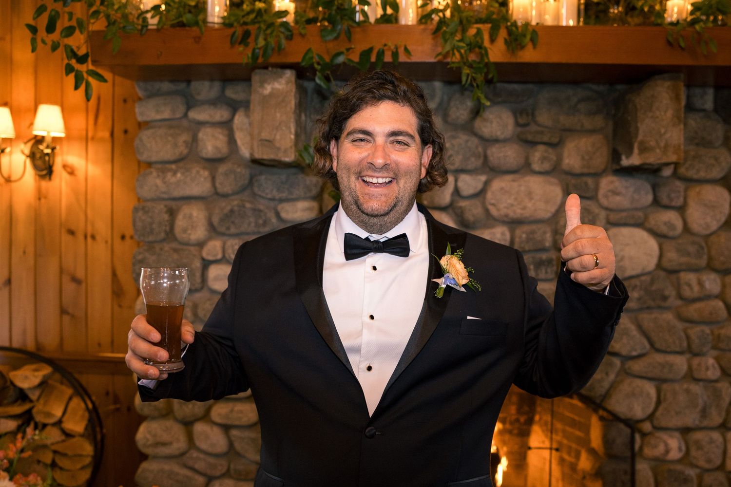 Groom in black tuxedo giving thumbs up while holding champagne glass in front of stone fireplace.