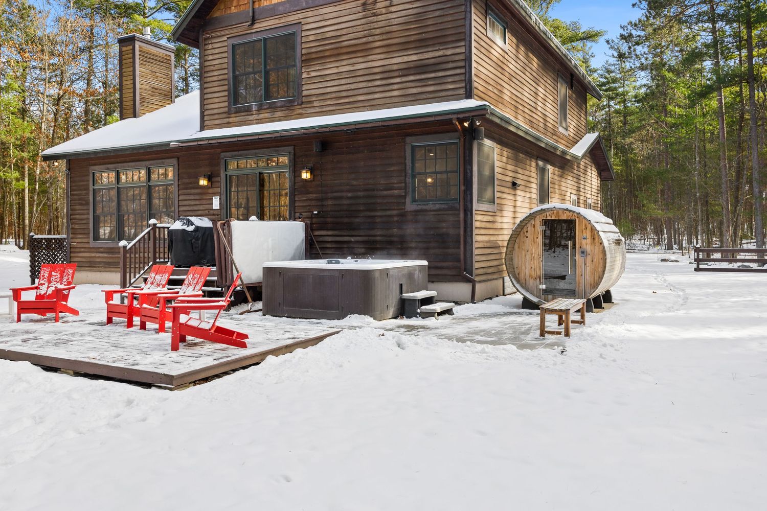 Modern wooden cabin with large windows nestled in a snowy forest setting, featuring red outdoor furniture and rustic charm.