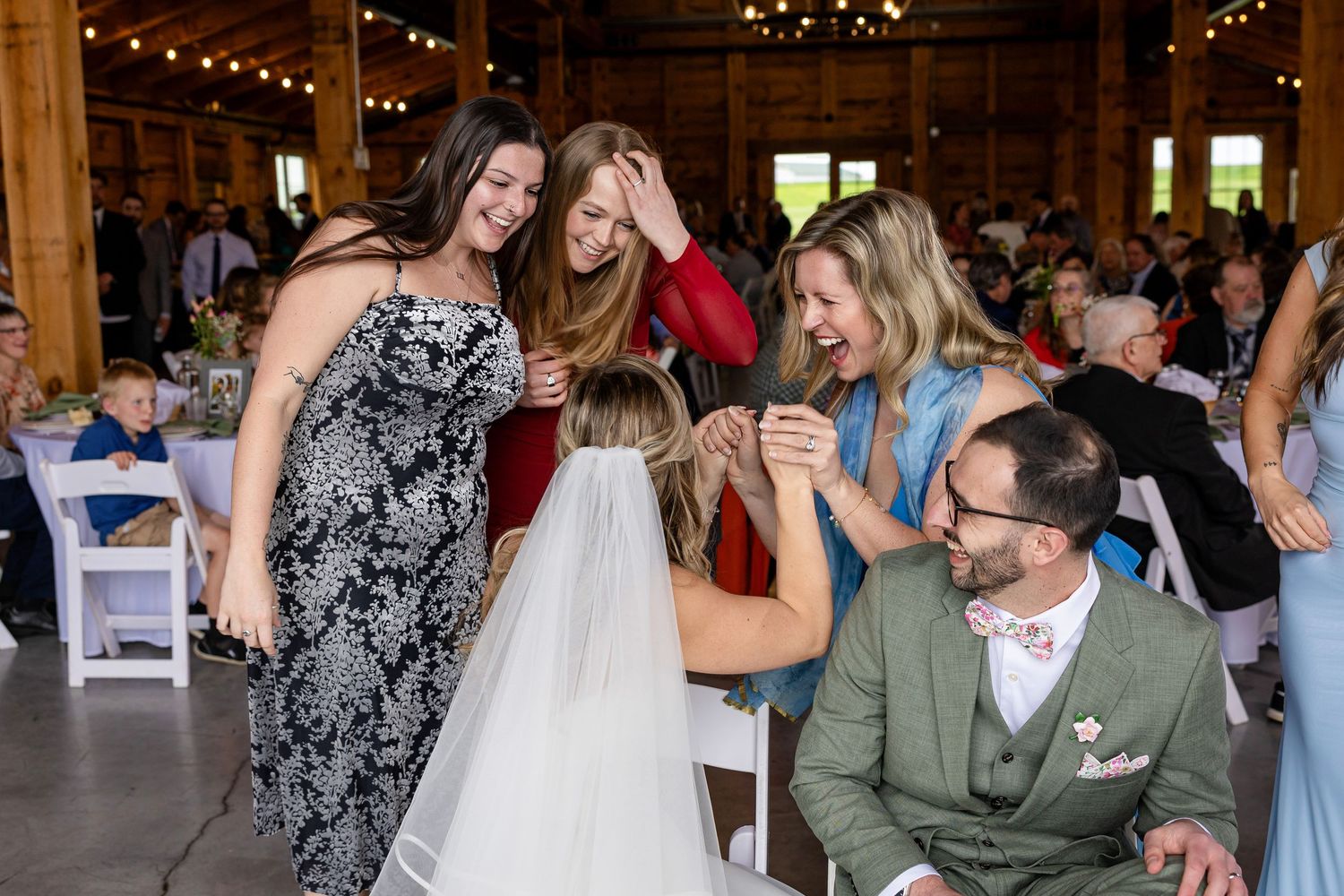 Joyful wedding guests dancing and celebrating with bride in elegant barn venue decorated with string lights.