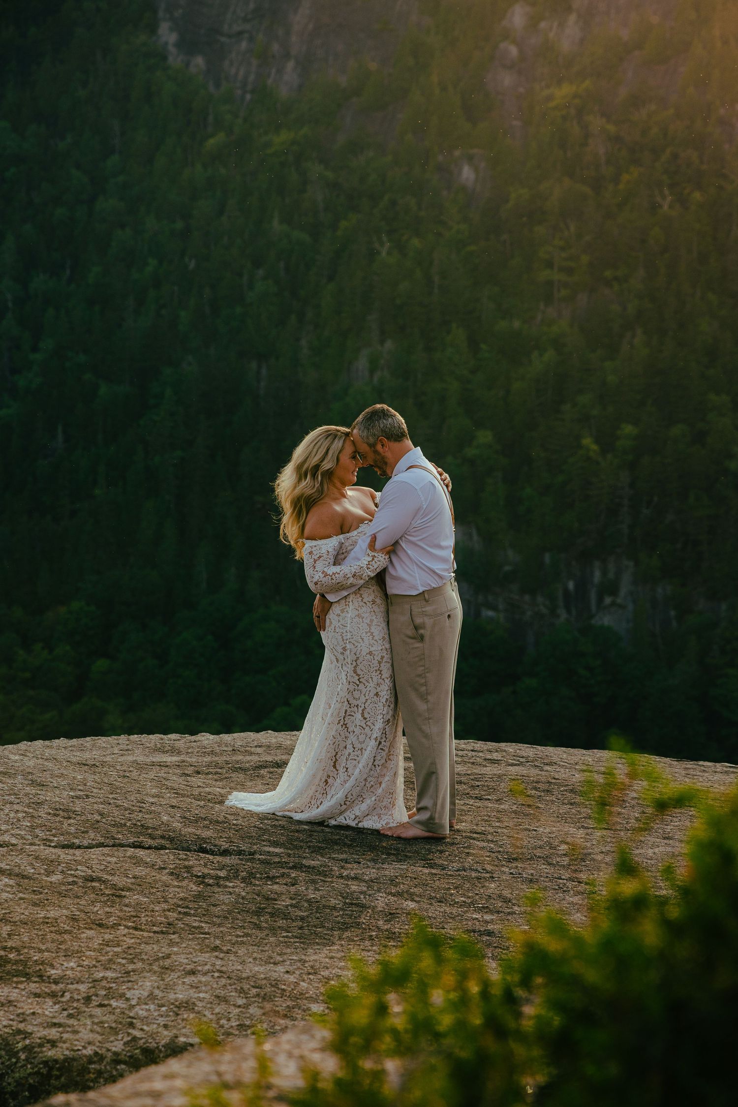 A couple shares an intimate moment at sunset on a rocky mountain overlook with forest in the background.