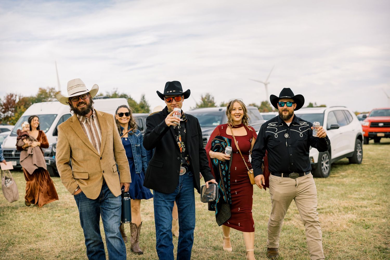 Group of five people in western wear and cowboy hats walking across a grassy field with parked cars and trees in the background.