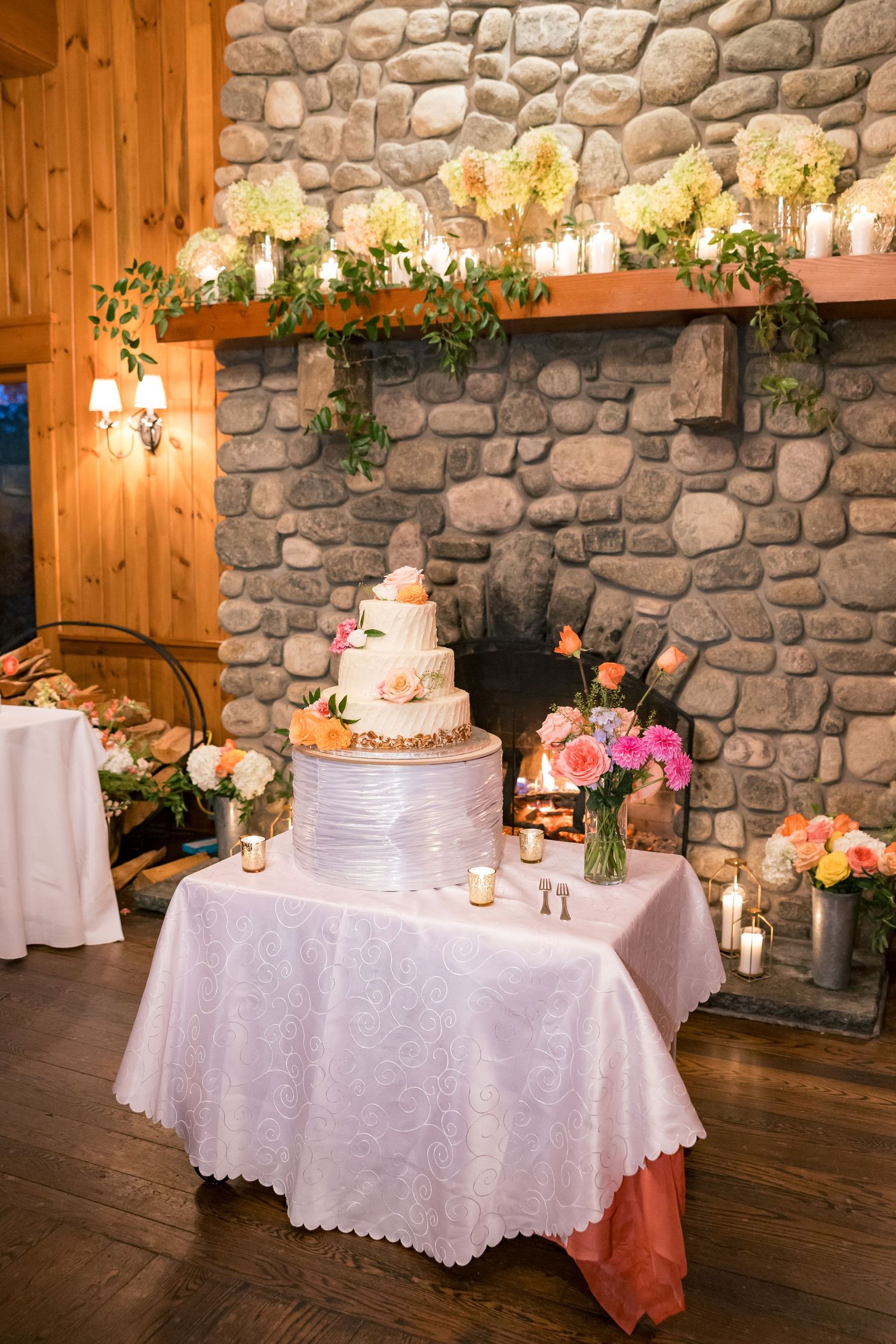 Elegant wedding cake display on lace-covered table before stone fireplace, adorned with pink roses and white floral mantel decor.