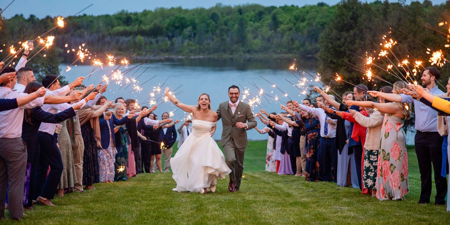 Bride and groom walk through sparkler tunnel surrounded by wedding guests near a scenic lakeside venue.