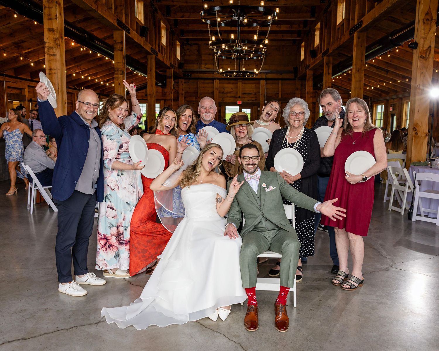 Joyful wedding group photo with bride and groom surrounded by family and friends in a rustic barn venue.
