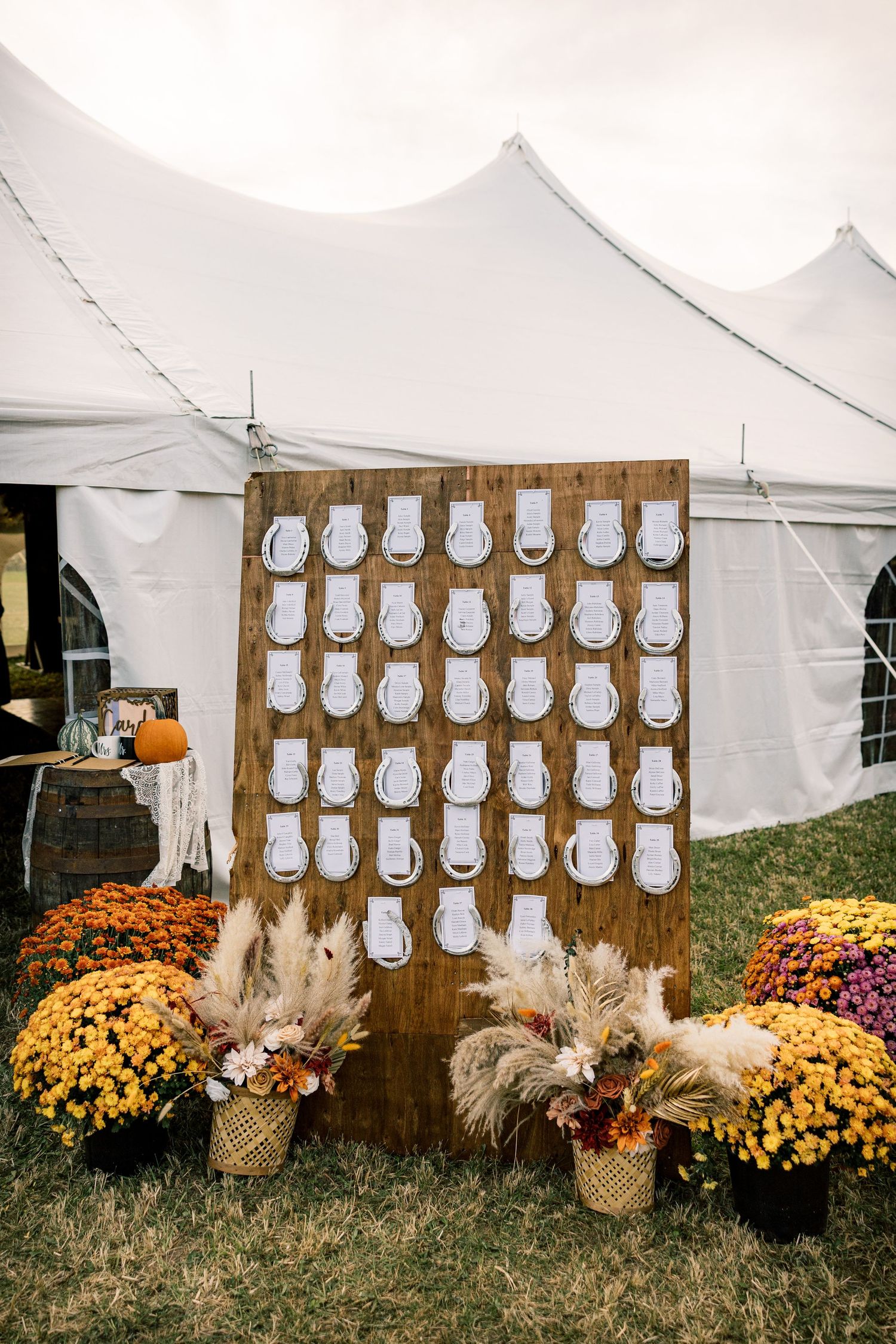 Rustic wooden wedding seating chart with horseshoe table cards beside fall mums and pampas grass in front of a white tent.