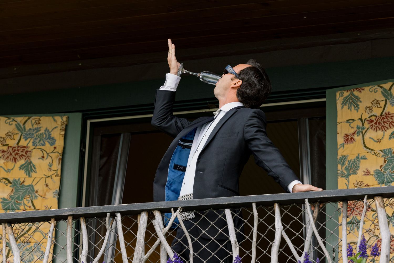 Man in suit drinking from bottle on balcony with floral wallpaper background.