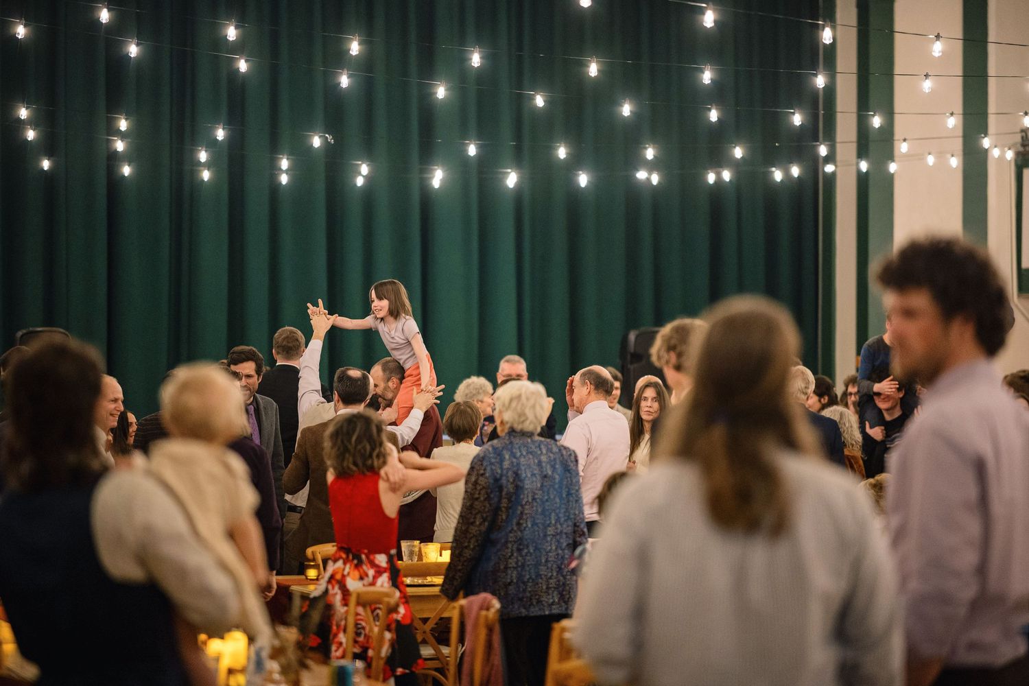 Wedding guests dance and celebrate under twinkling string lights at an indoor reception venue.