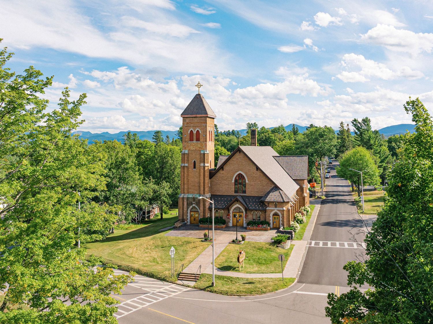 Aerial view of a historic brick church beside a tree-lined small town street with mountain views under a partly cloudy sky.