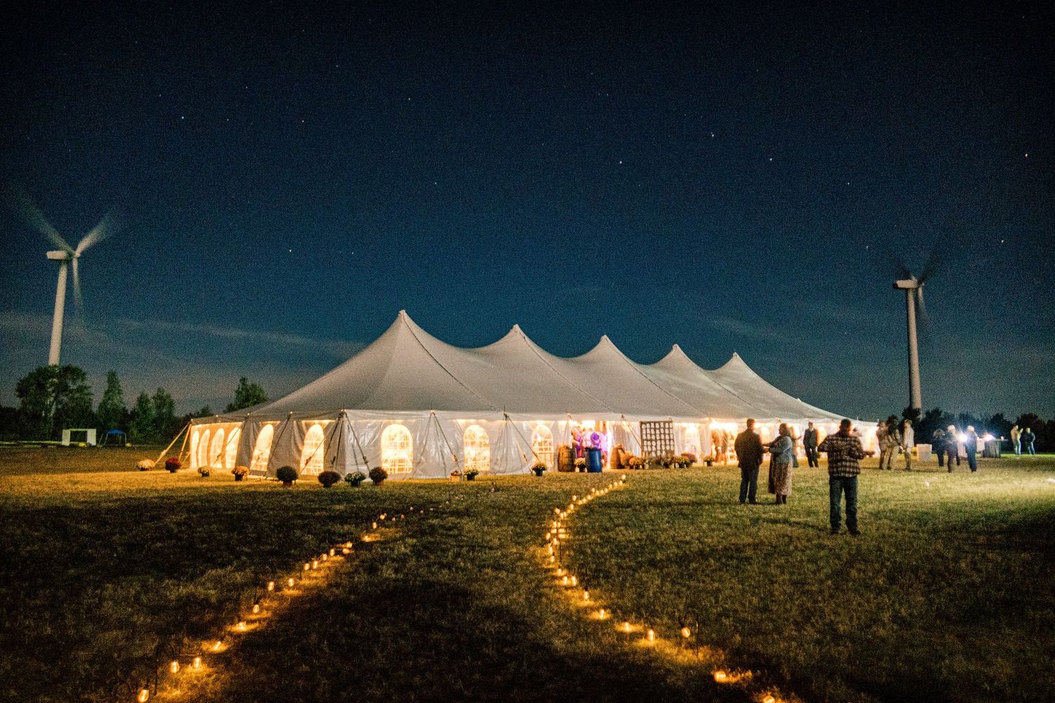 Illuminated white wedding tent glowing under a starry night sky, with lantern-lit pathways and wind turbines in the background.
