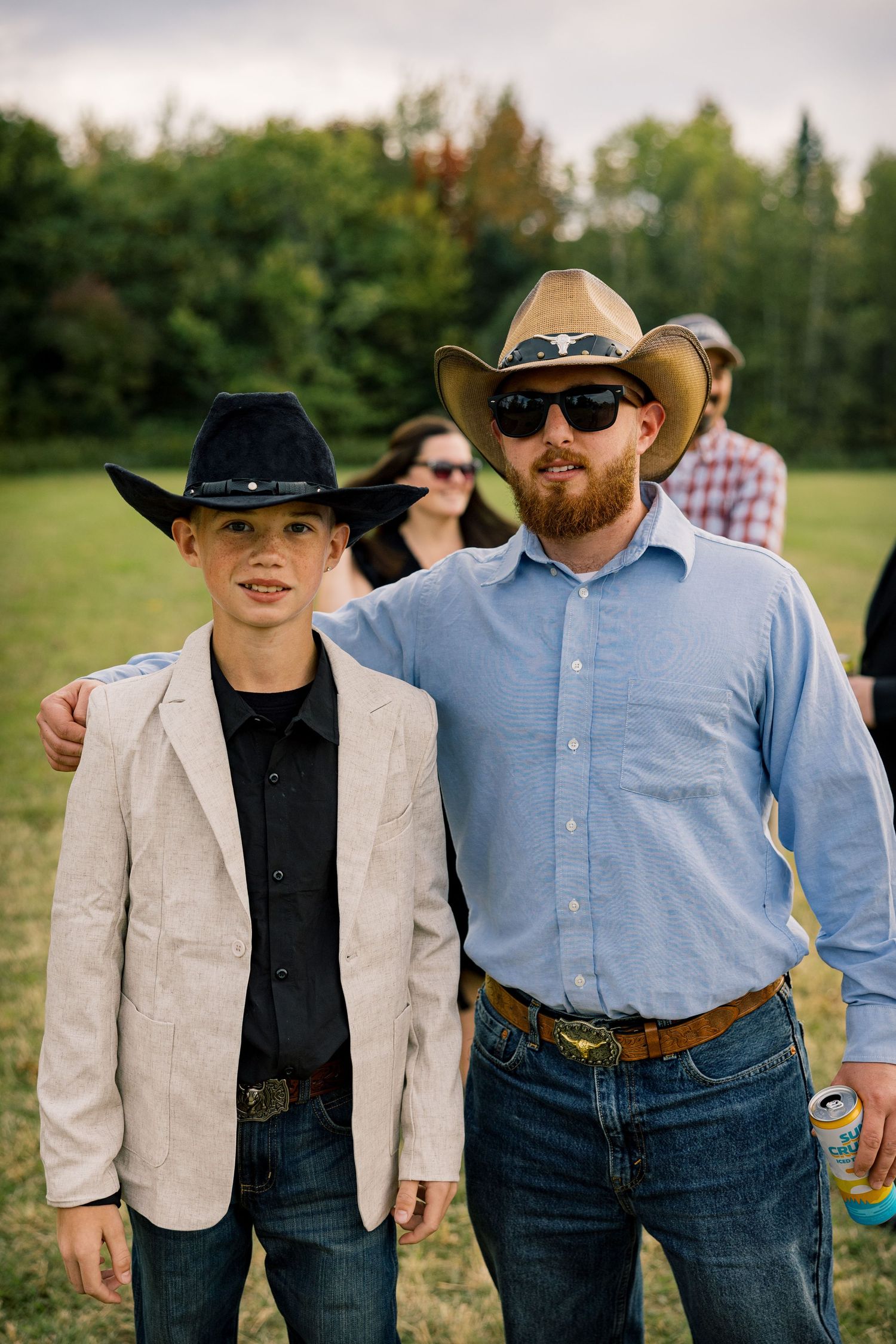 A bearded man in a straw cowboy hat and sunglasses poses with a boy in a black cowboy hat on a grassy field.
