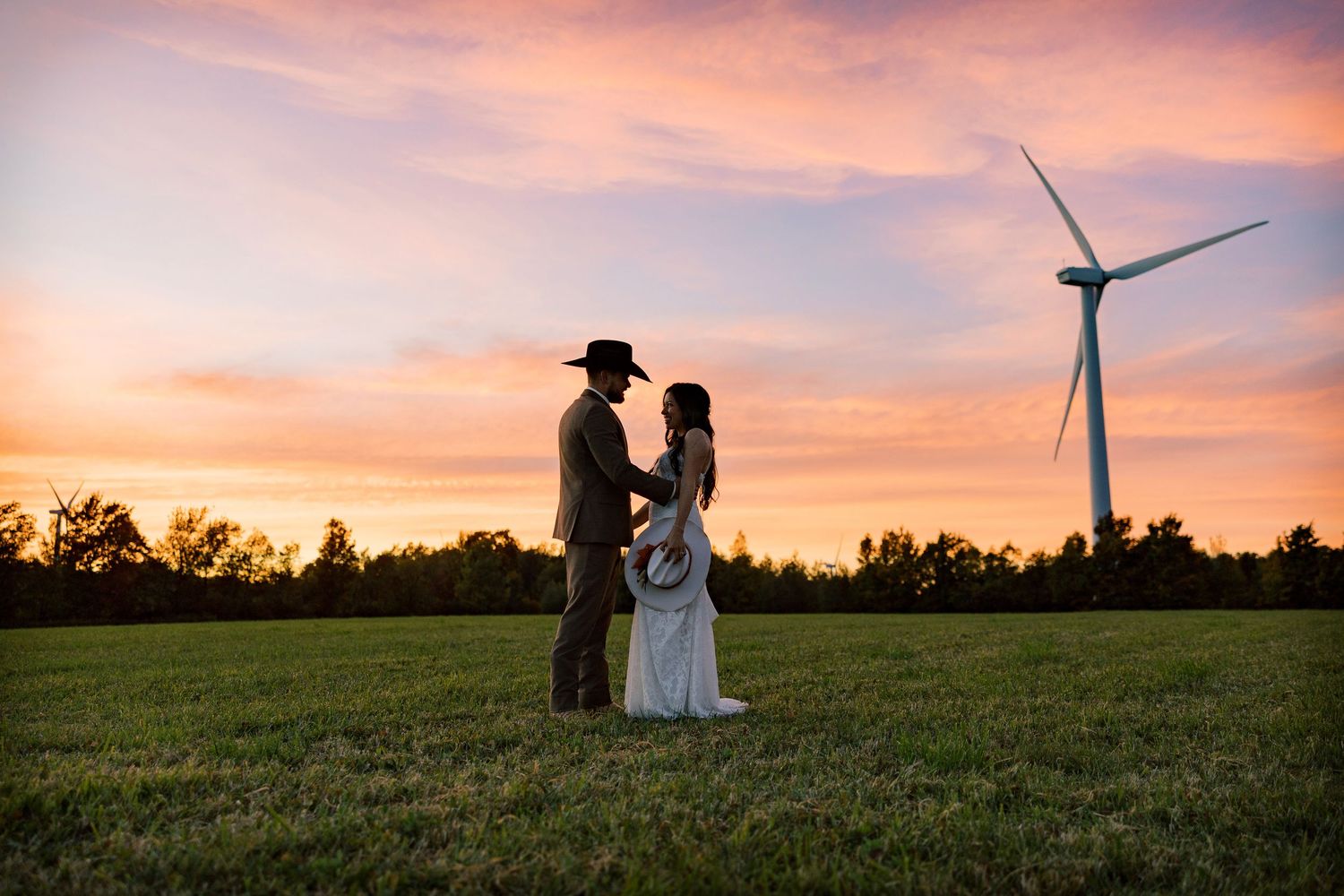 Bride in a white gown and groom in a cowboy hat embrace in a grassy field at sunset with a wind turbine and pink sky behind them.