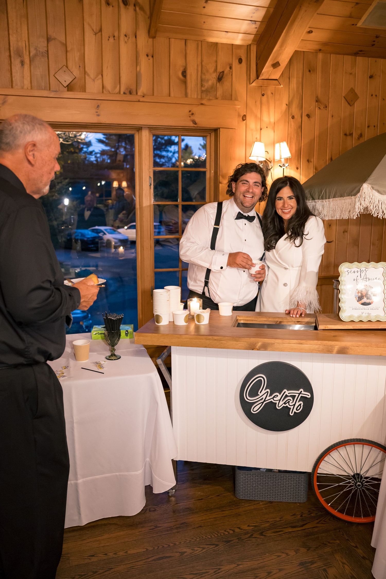 Two smiling gelato cart servers in bow ties assist a guest at an elegant indoor wedding reception venue.