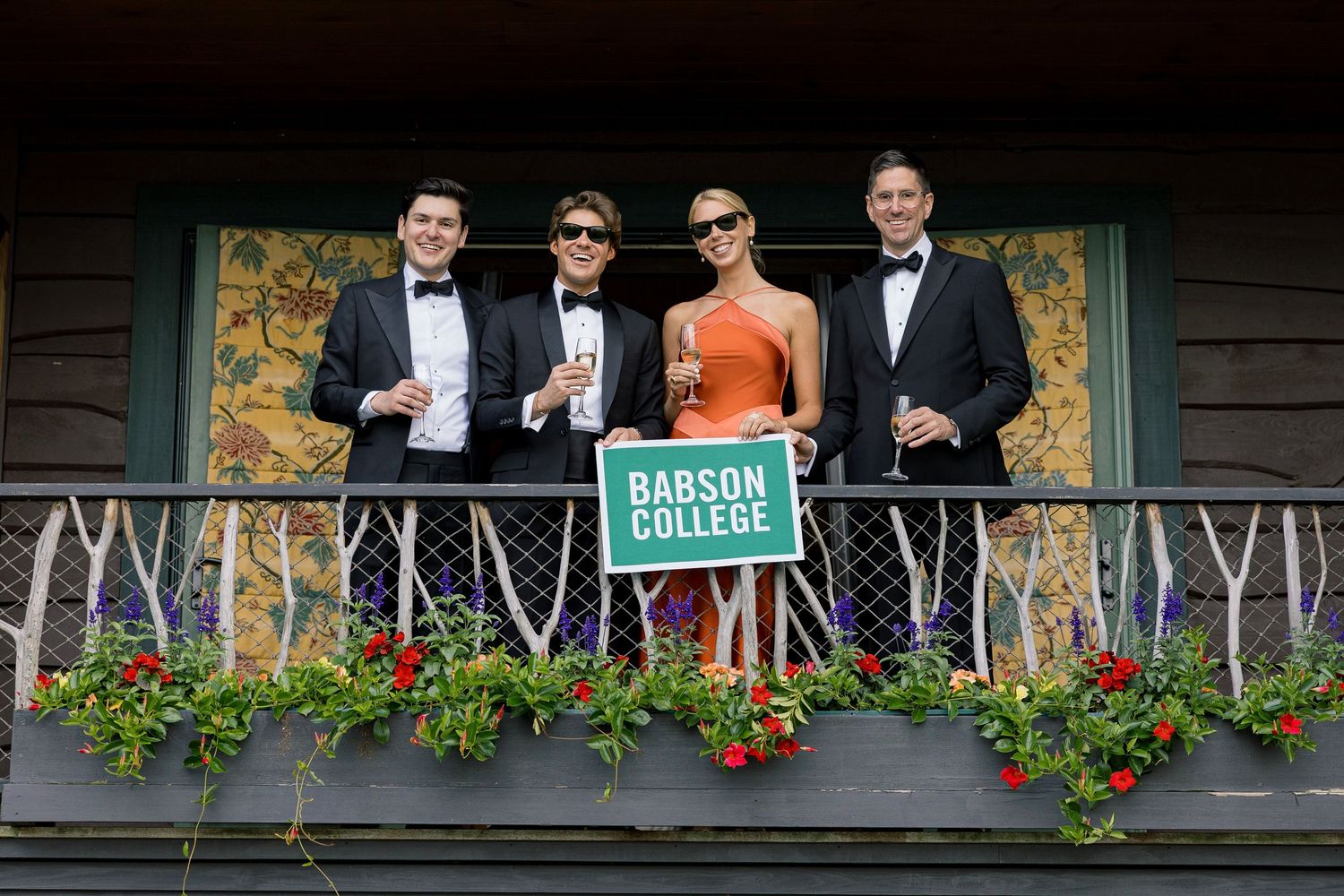 Four formally dressed students pose on a balcony with a Babson College sign surrounded by colorful flowers.