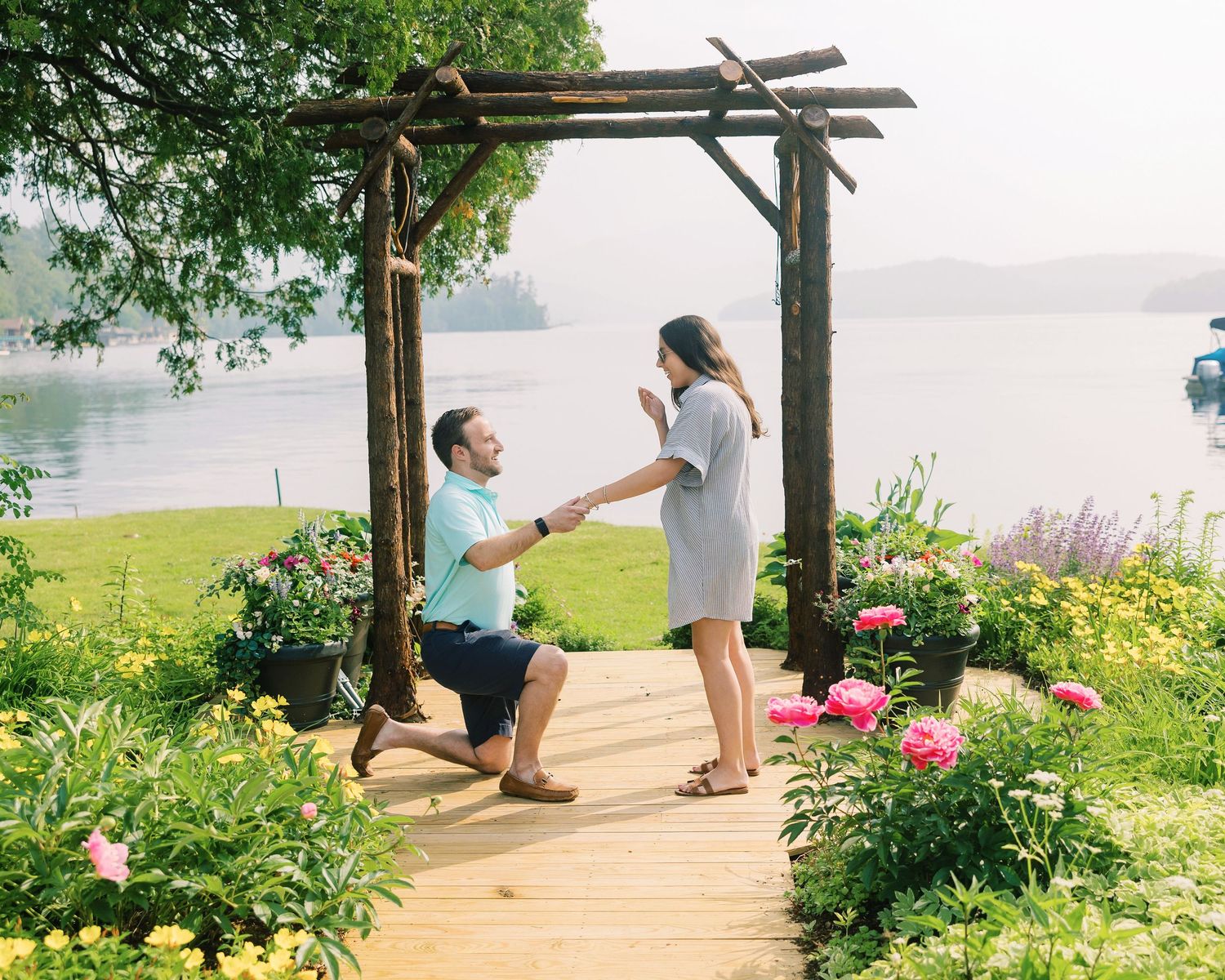 A proposal moment under a wooden pergola surrounded by colorful flowers near a lake.