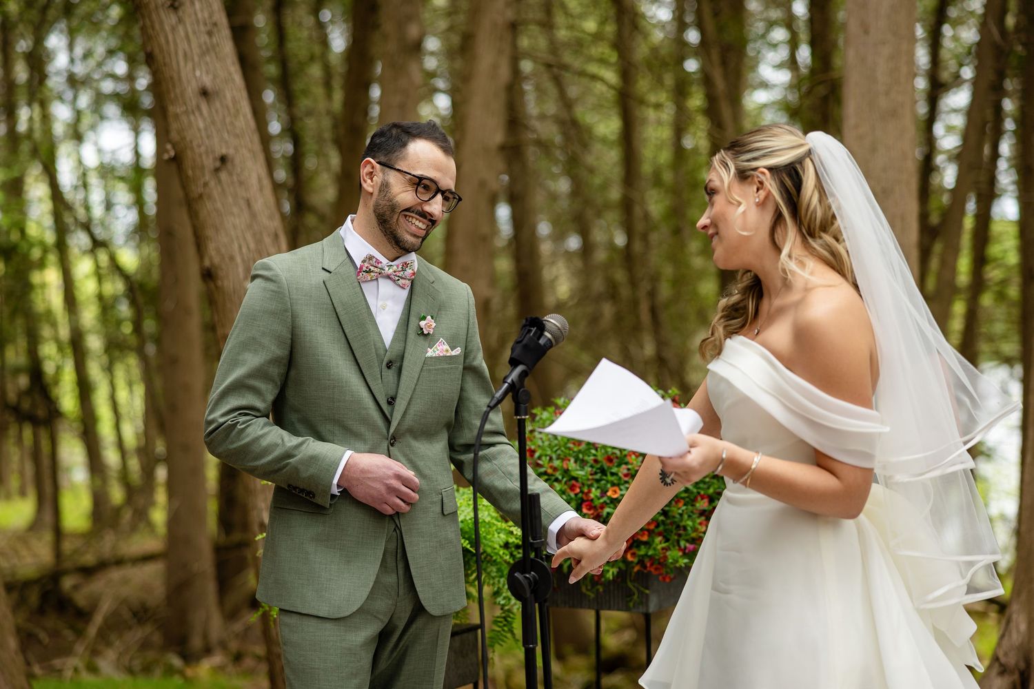 Bride reading vows to groom in sage green suit during outdoor forest wedding ceremony.