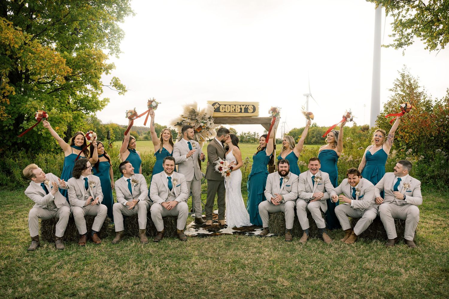 Bride and groom pose outdoors with bridesmaids in teal dresses raising bouquets and groomsmen in tan suits seated on the grass.