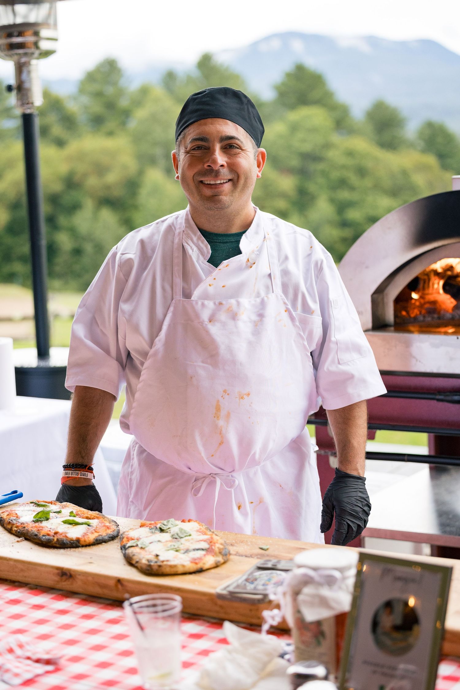 Smiling chef in white uniform stands beside fresh Neapolitan pizzas at an outdoor wood-fired oven.