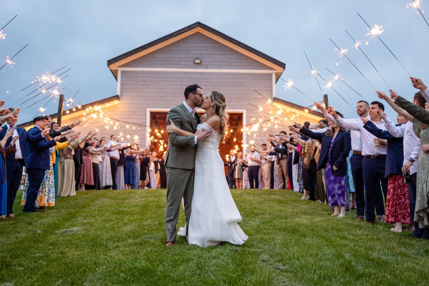 Bride and groom share a kiss during a sparkler send-off at dusk outside a white barn wedding venue.