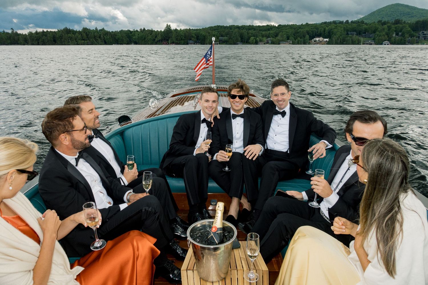 Wedding party in tuxedos celebrating on a classic wooden boat on a lake, toasting with champagne glasses.