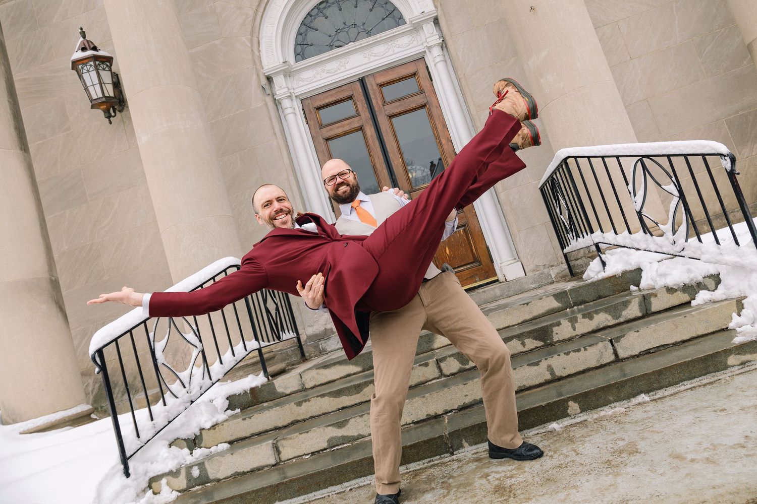 A ballet dancer in a red coat performs an elegant arabesque pose on snowy stone steps.