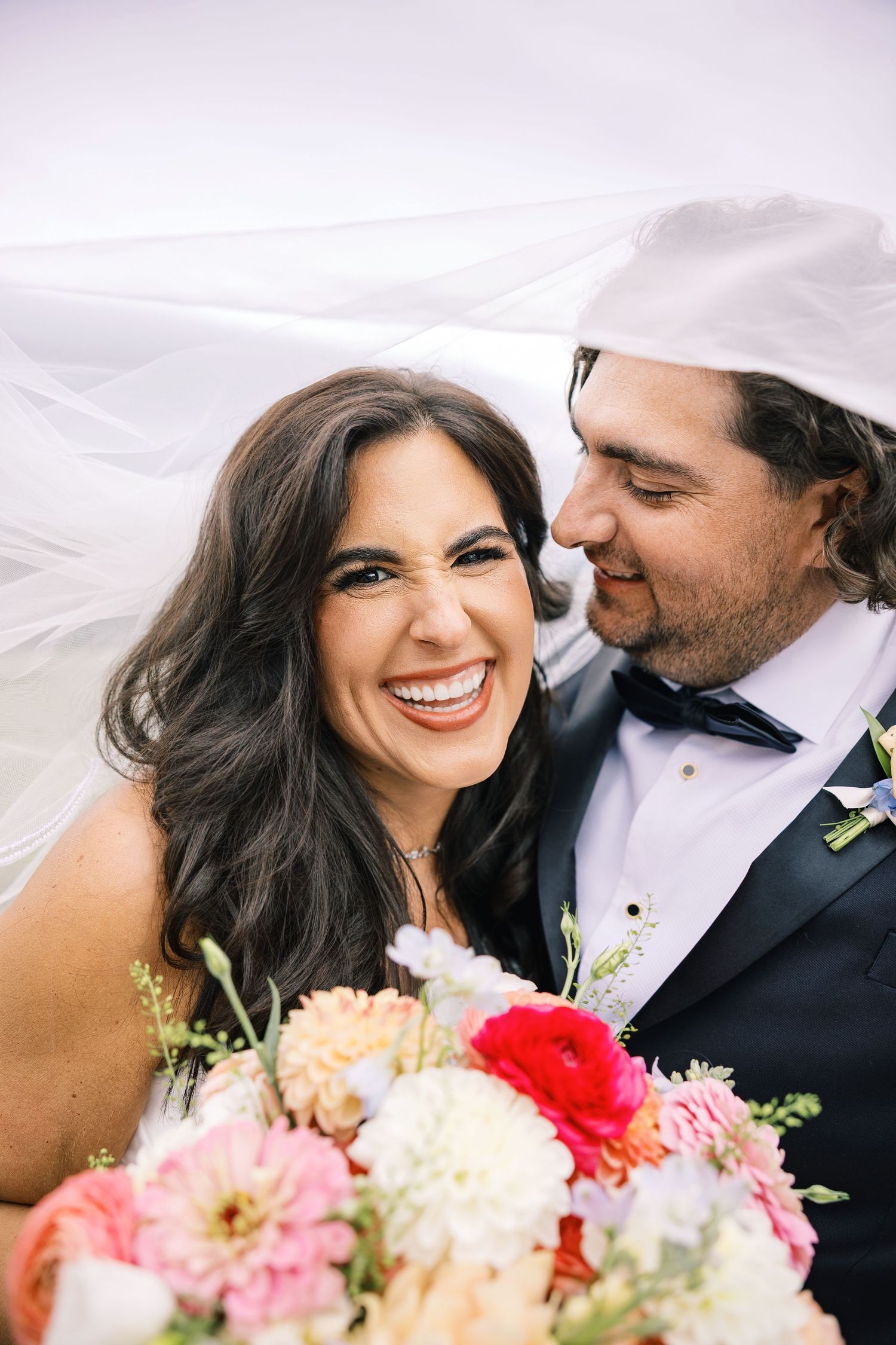 Smiling bride with dark hair holds colorful bouquet beside groom in tuxedo under flowing wedding veil.