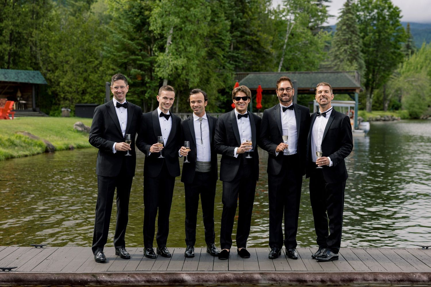 Six groomsmen in black tuxedos holding champagne glasses on a dock by a scenic lake surrounded by lush green trees.