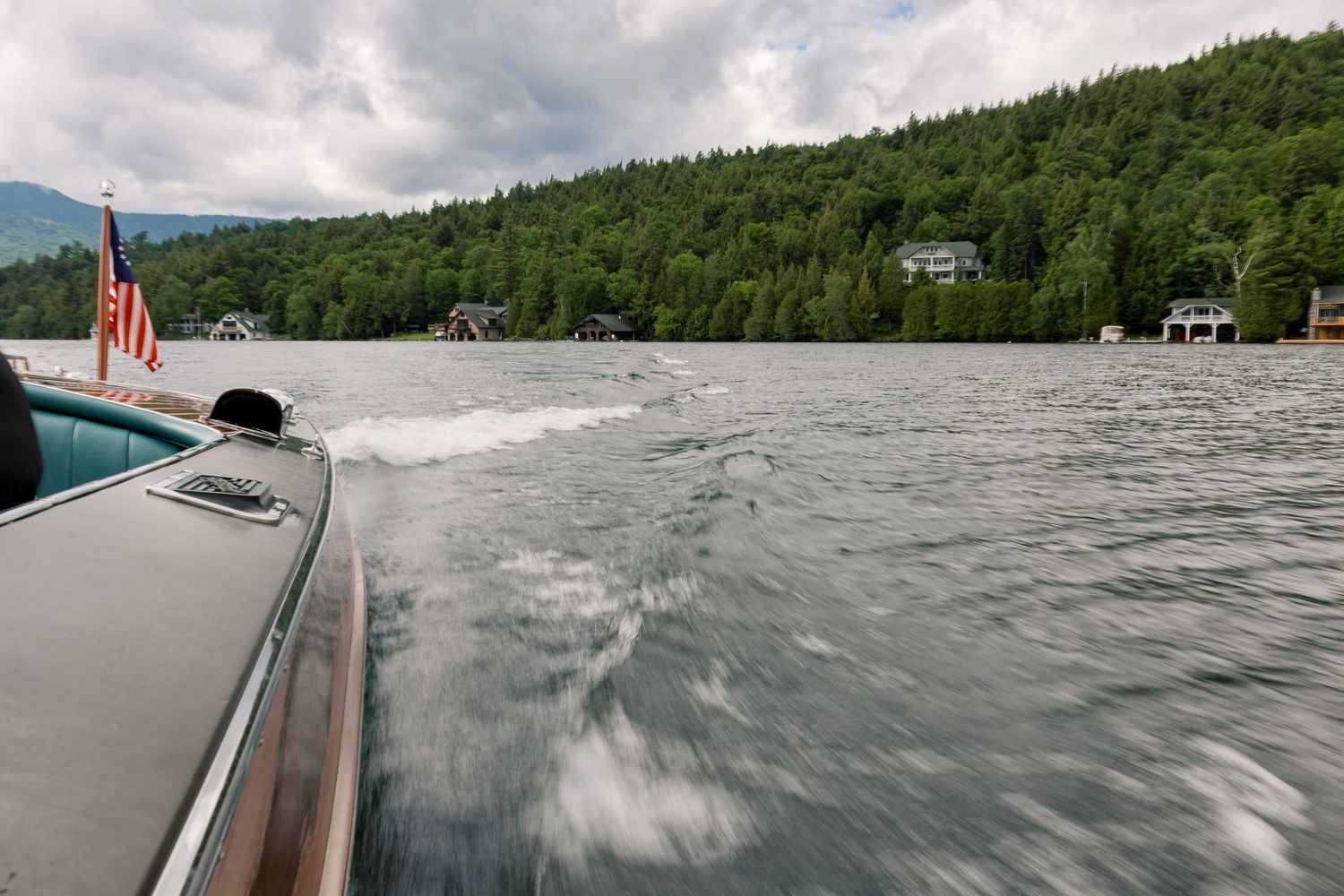 View from a motorboat on a calm lake with forested hills, lakeside homes, and an American flag in the background.