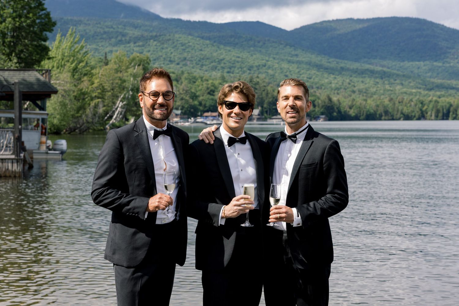Three men in tuxedos holding champagne glasses smile by a scenic mountain lake on a sunny day.