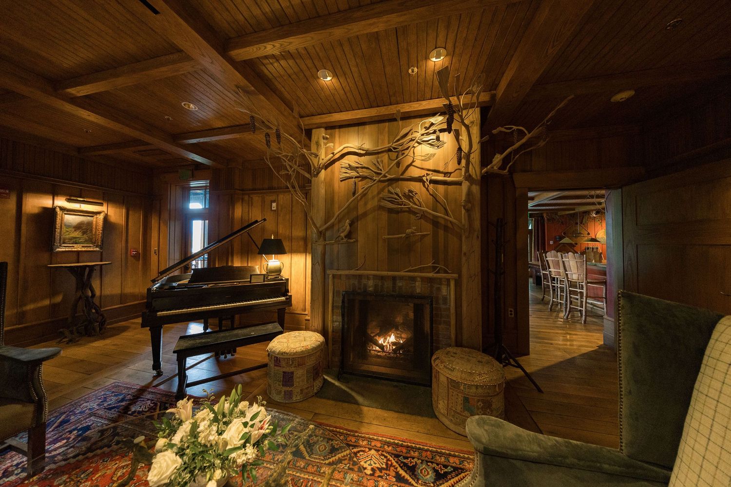 Cozy rustic wood-paneled living room with a grand piano, ornate stone fireplace, and warm glowing fire.