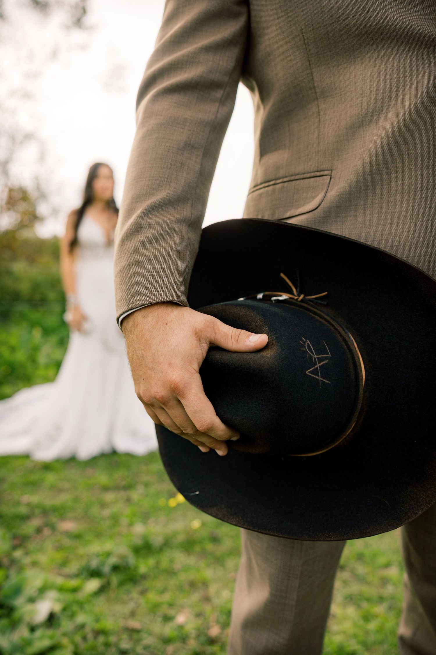 Groom in a tan suit holds a black cowboy hat with initials while a bride in a white wedding gown stands blurred on the grass behind him.