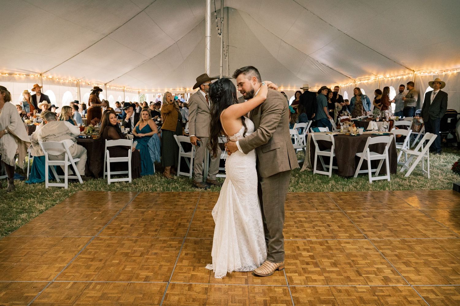 Bride in a white lace gown and groom in a gray suit share a first dance on a wooden dance floor under a wedding tent as guests look on.