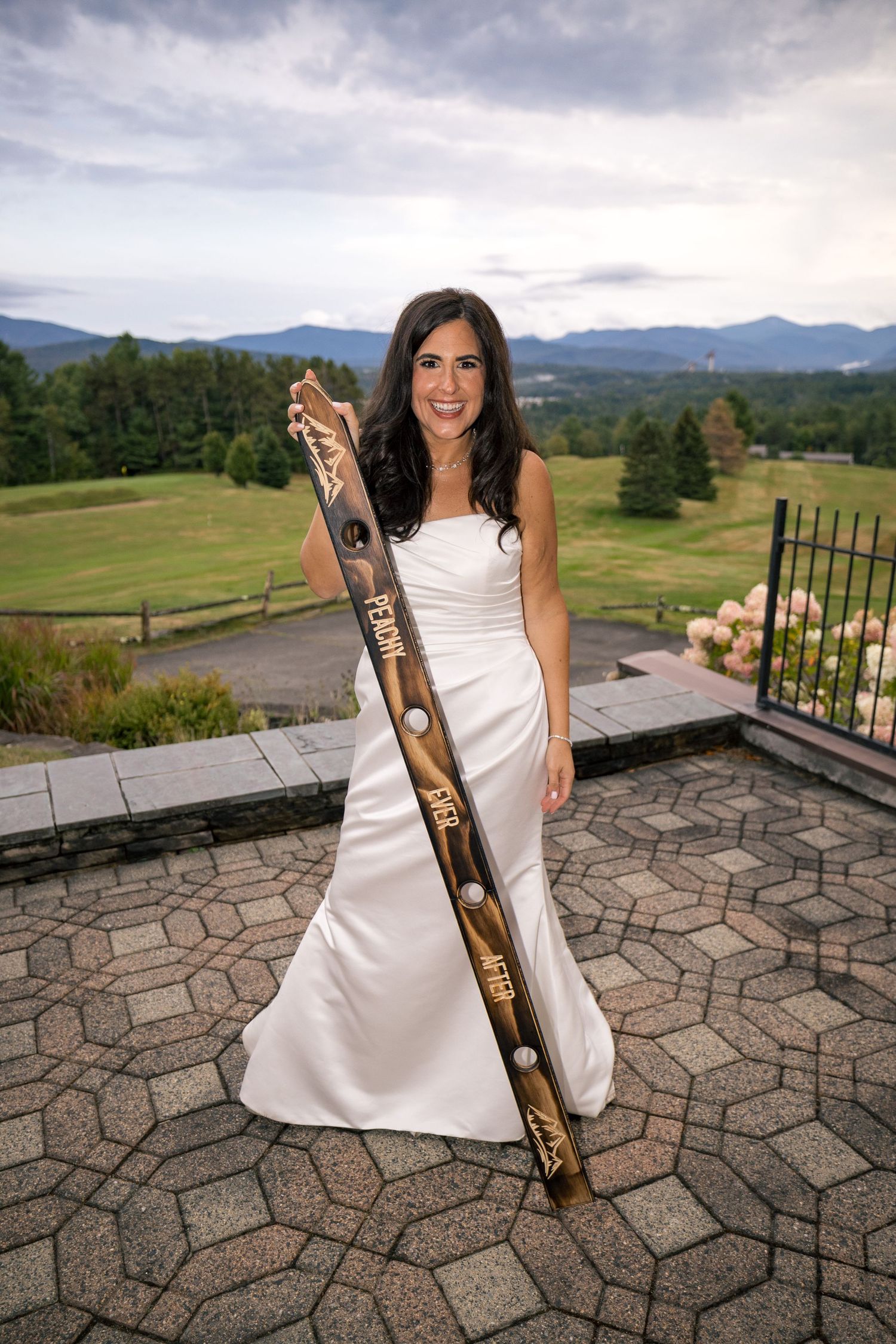 Bride in white gown holding decorated ski on stone terrace with mountain golf course backdrop.
