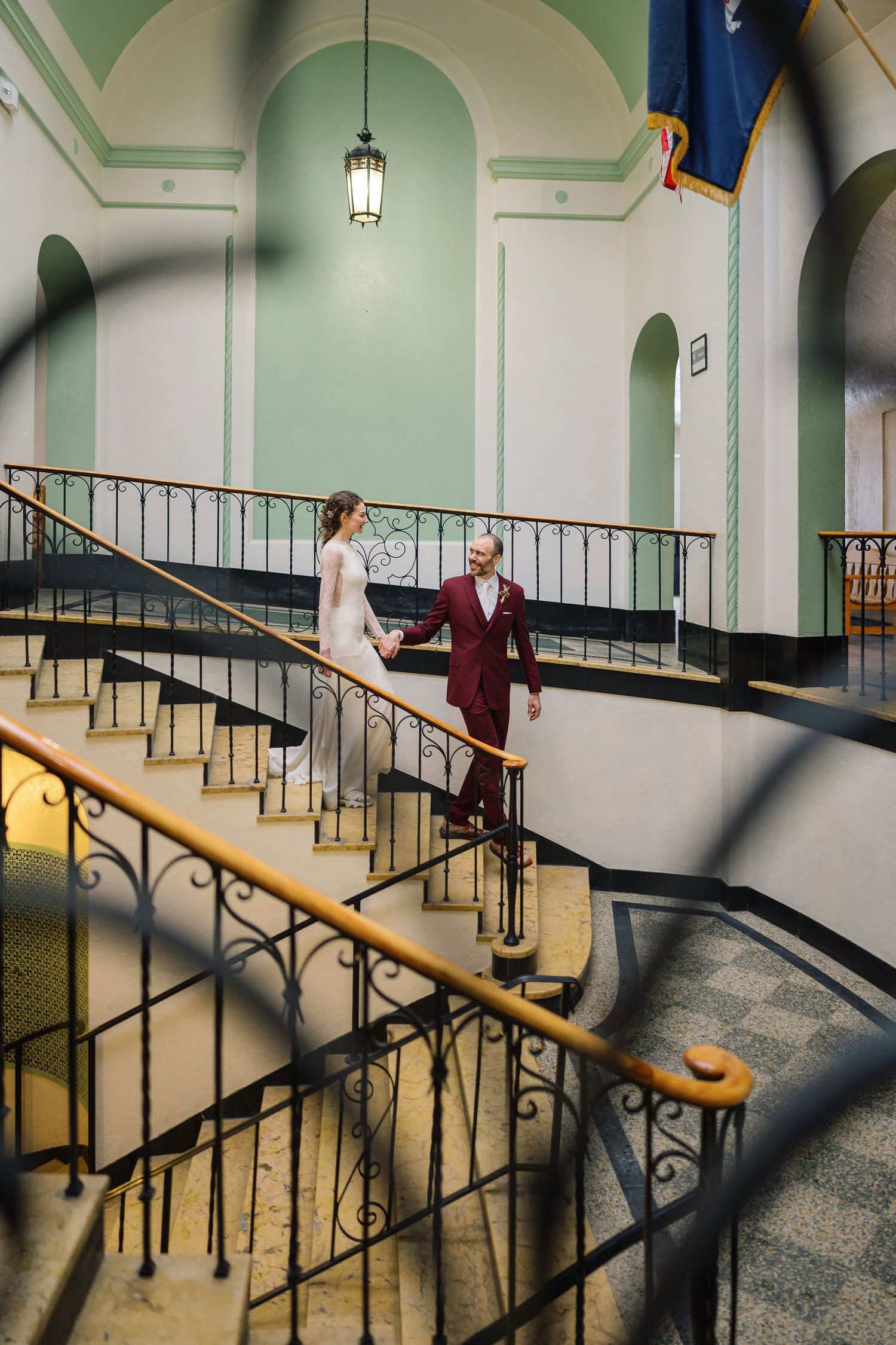 A person in a white coat walks up a vintage staircase with ornate railings and mint green walls in a hospital setting.